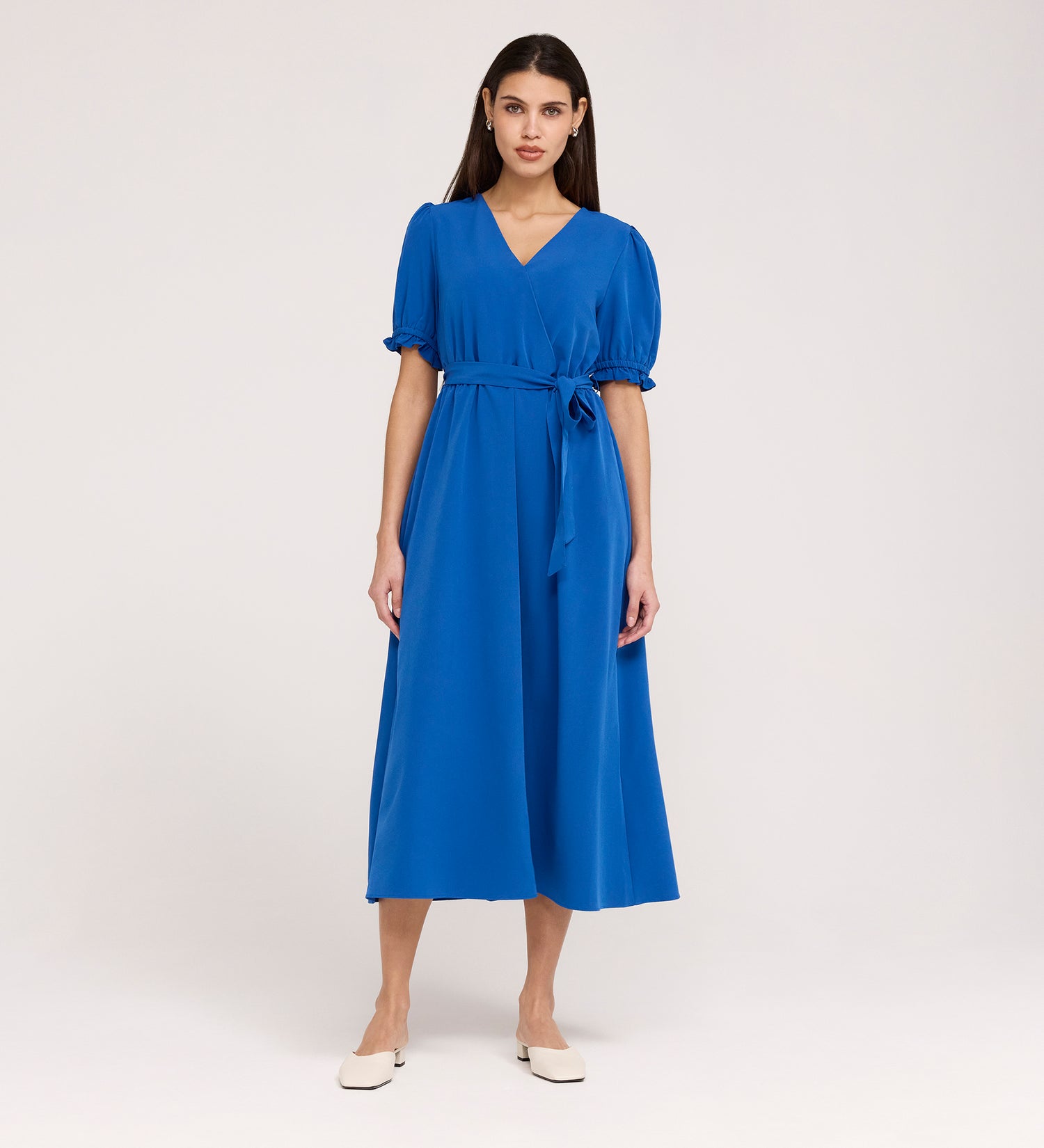 A woman stands against a plain light background wearing the Everleigh Cobalt Crepe Midi Dress—a bright blue, short-sleeve, faux wrap midi with waist tie—paired with white flats.