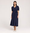 A woman stands against a plain background wearing the Everleigh Navy Crepe Midi Dress—a navy, short-sleeved midi with a faux wrap neckline, tied waist, and white flat shoes.