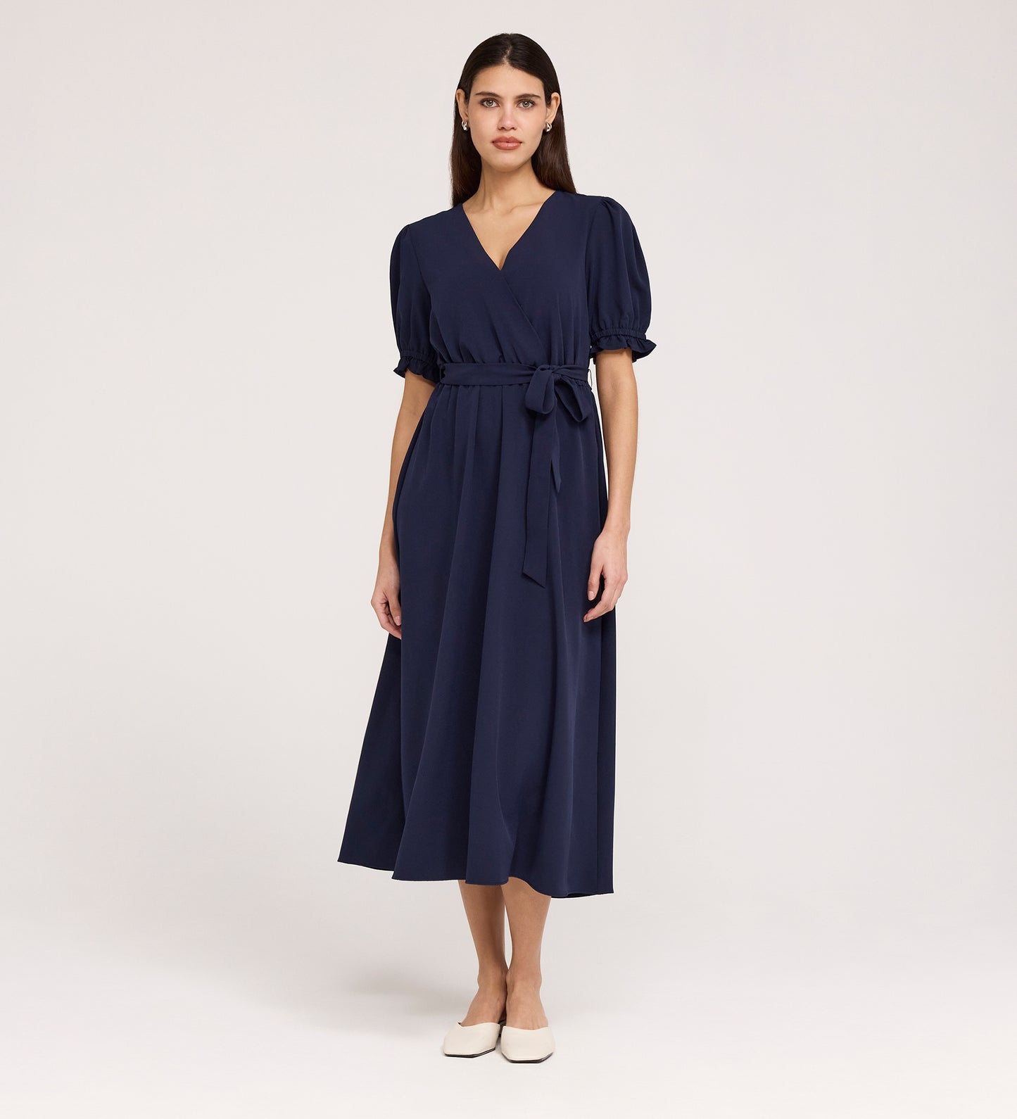 A woman stands against a plain background wearing the Everleigh Navy Crepe Midi Dress—a navy, short-sleeved midi with a faux wrap neckline, tied waist, and white flat shoes.