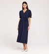 A woman with straight, dark hair and a neutral expression wears the Everleigh Navy Crepe Midi Dress—featuring short sleeves, a faux wrap neckline, tied waist—against a plain background. She pairs it with white flat shoes.