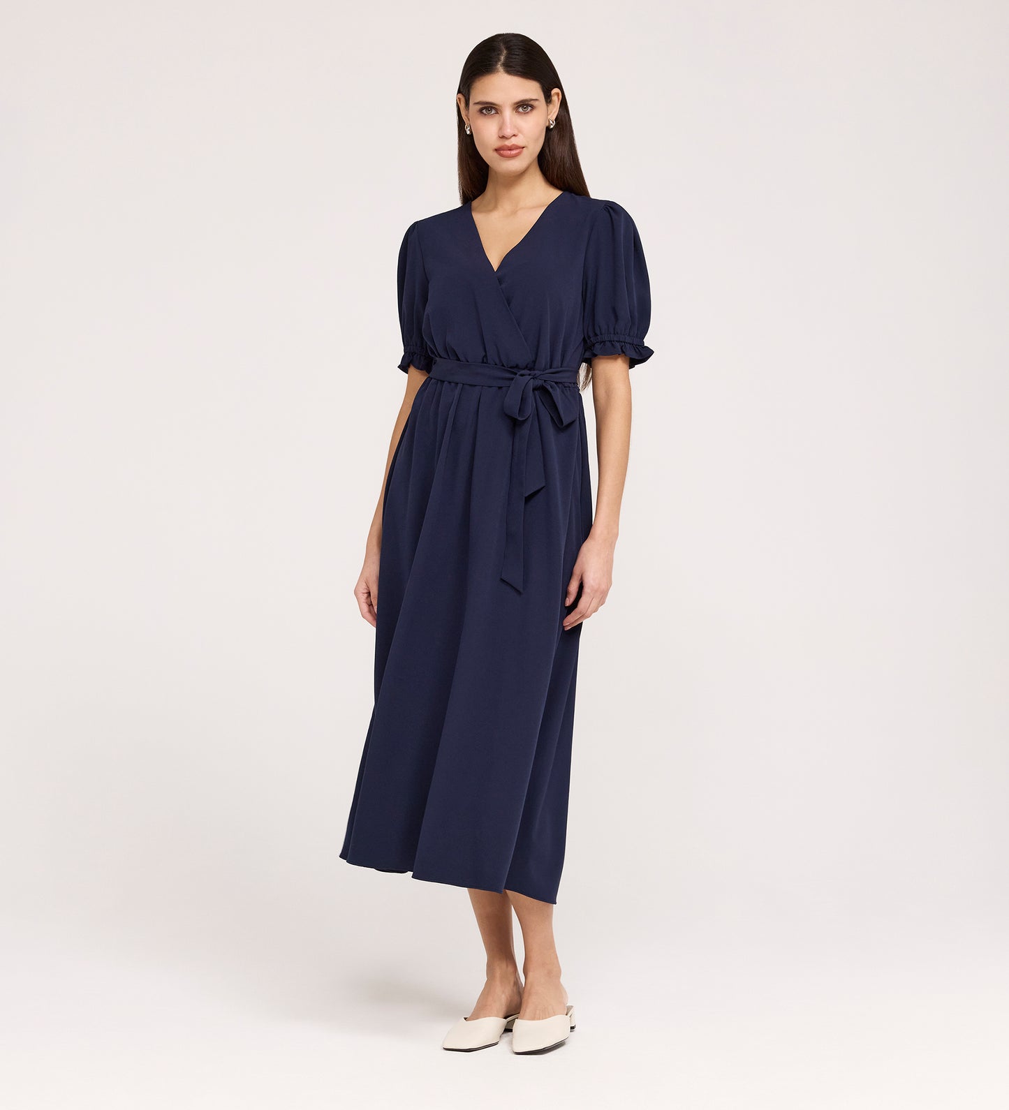 A woman with straight, dark hair and a neutral expression wears the Everleigh Navy Crepe Midi Dress—featuring short sleeves, a faux wrap neckline, tied waist—against a plain background. She pairs it with white flat shoes.