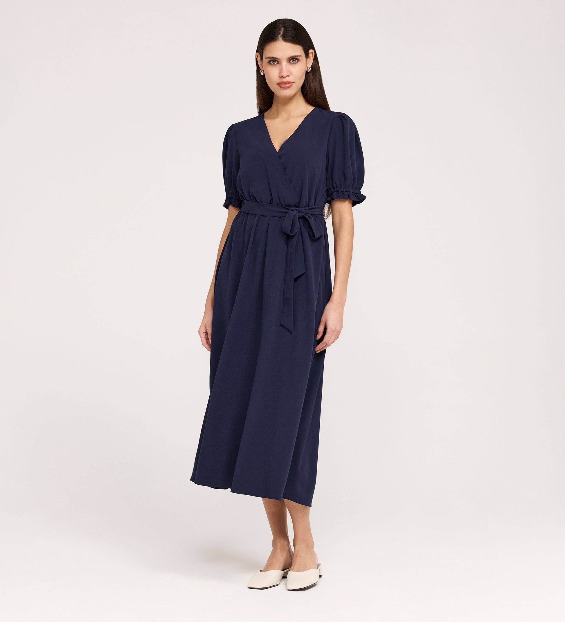 A woman with straight, dark hair and a neutral expression wears the Everleigh Navy Crepe Midi Dress—featuring short sleeves, a faux wrap neckline, tied waist—against a plain background. She pairs it with white flat shoes.