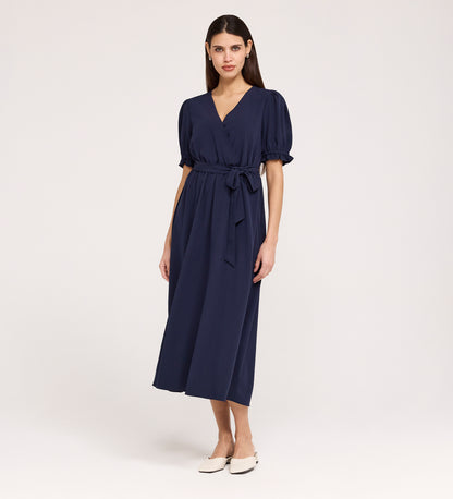 A woman with straight, dark hair and a neutral expression wears the Everleigh Navy Crepe Midi Dress—featuring short sleeves, a faux wrap neckline, tied waist—against a plain background. She pairs it with white flat shoes.