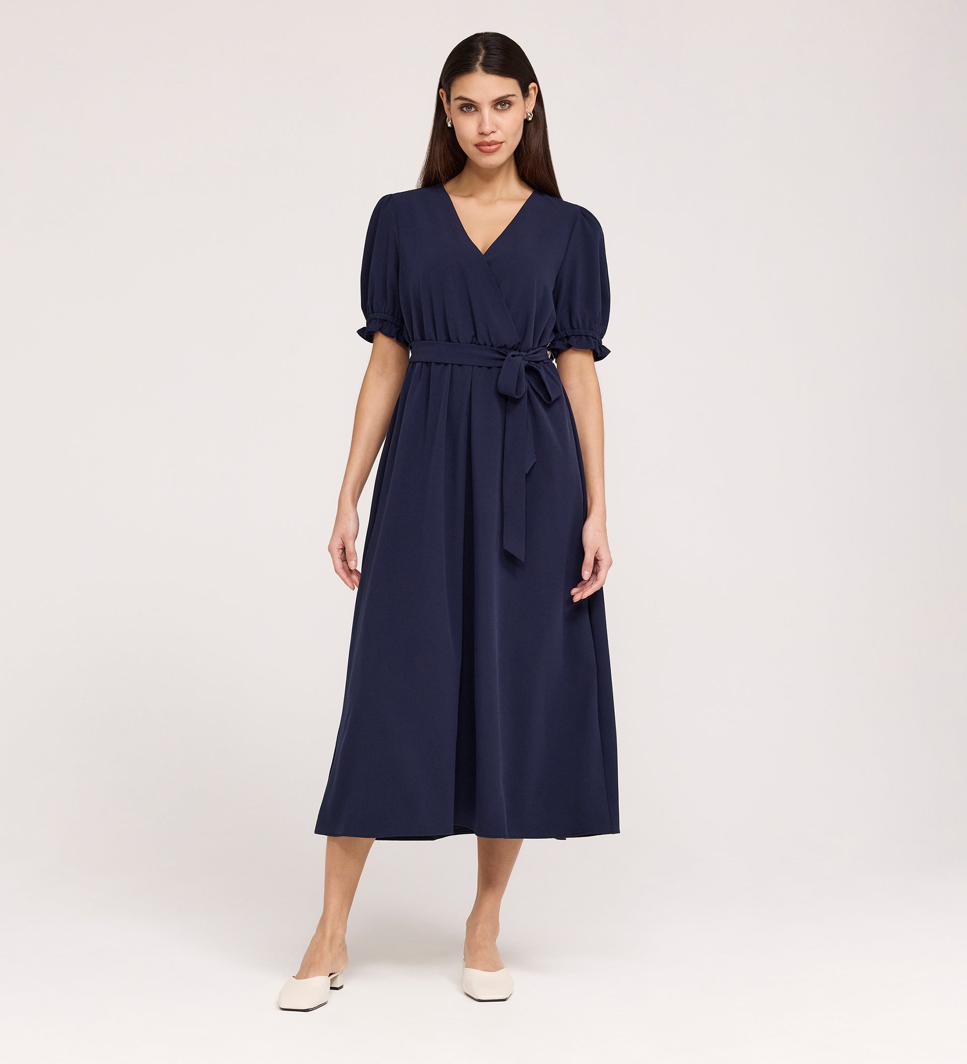A woman with long, straight dark hair stands against a light background wearing the Everleigh Navy Crepe Midi Dress— a short-sleeve, belted midi dress with a faux wrap neckline— paired with beige flat shoes.