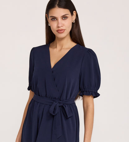 Woman in the Everleigh Navy Crepe Midi Dress—featuring short puff sleeves, a faux wrap neckline, and a waist tie—stands against a plain light background.