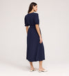 A woman with long brown hair, seen from behind, wears the Everleigh Navy Crepe Midi Dress featuring a faux wrap neckline and elastic waist. She pairs it with cream flats and stands against a plain light background.