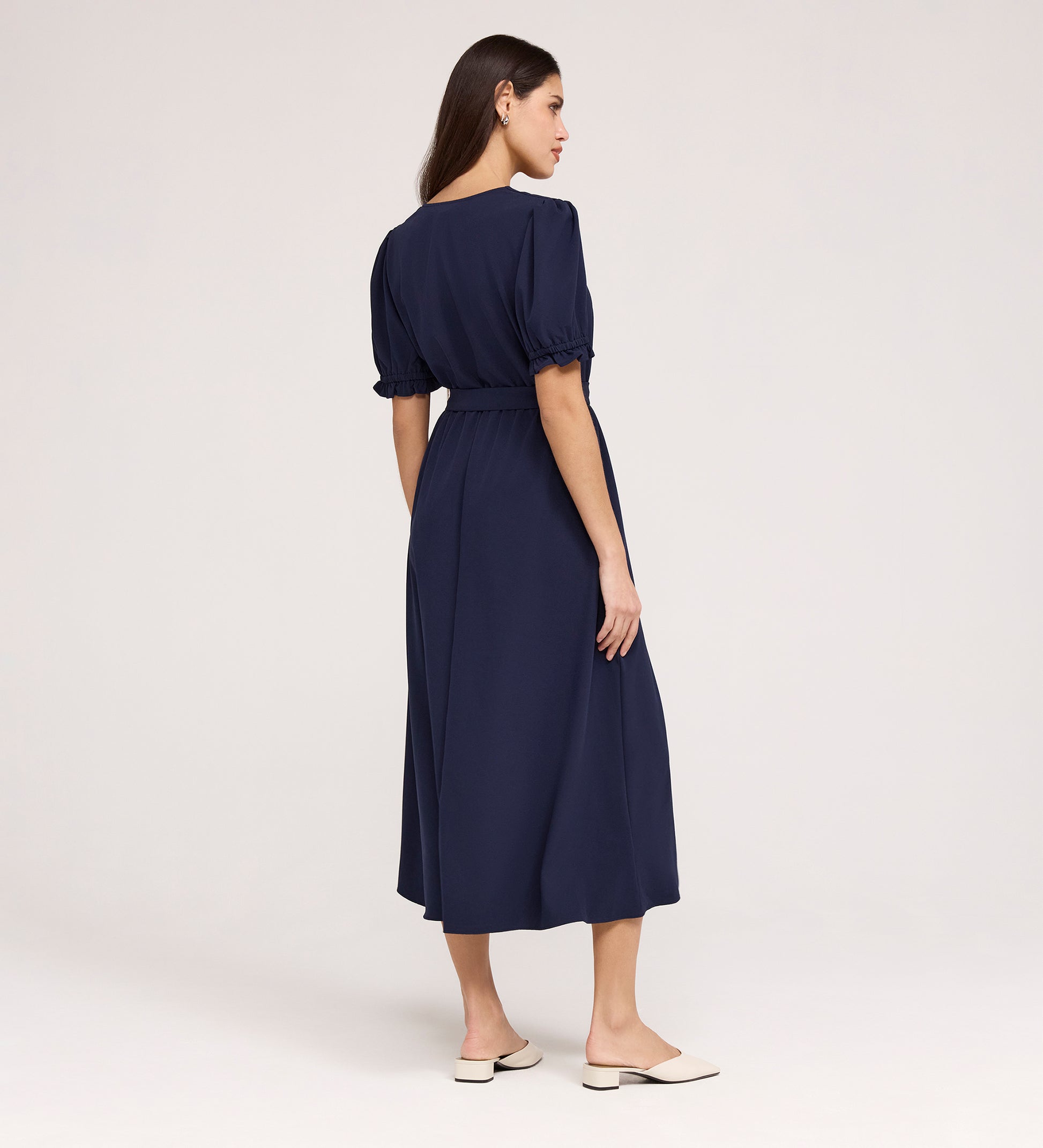 A woman with long brown hair, seen from behind, wears the Everleigh Navy Crepe Midi Dress featuring a faux wrap neckline and elastic waist. She pairs it with cream flats and stands against a plain light background.