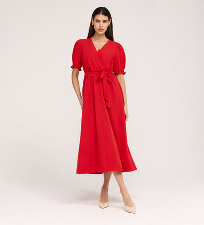 A woman stands against a plain background wearing the Everleigh Red Crepe Midi Dress, featuring a bright red hue, faux wrap neckline, puffed sleeves, waist tie, and nude pointed-toe heels.