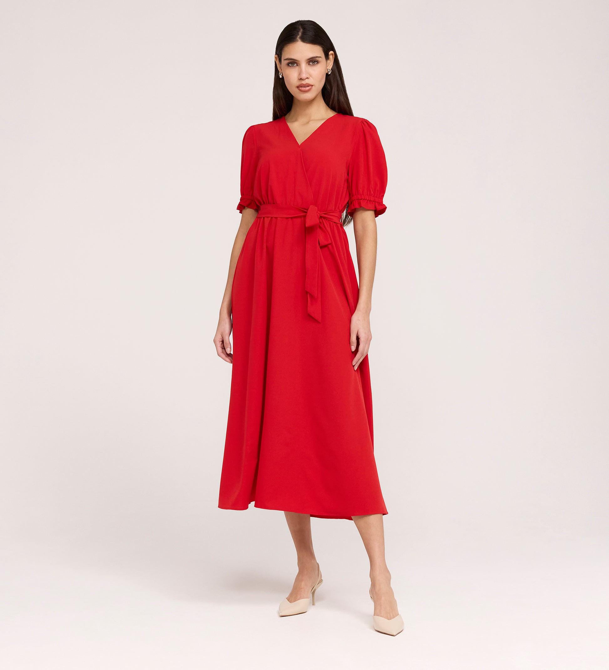 A woman stands in a studio, wearing the Everleigh Red Crepe Midi Dress—a versatile midi with a faux wrap neckline and tied waist belt—paired with beige pointed-toe heels against a plain light-colored background.