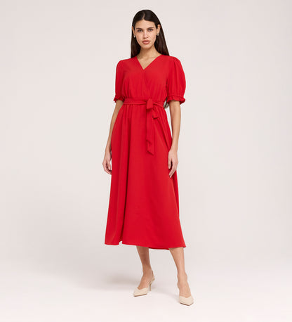 A woman stands in a studio, wearing the Everleigh Red Crepe Midi Dress—a versatile midi with a faux wrap neckline and tied waist belt—paired with beige pointed-toe heels against a plain light-colored background.
