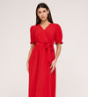 A woman with long dark hair wears the Everleigh Red Crepe Midi Dress—a bright red, short-sleeve midi with a tied waist and faux wrap neckline—standing against a plain light background.