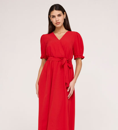 A woman with long dark hair wears the Everleigh Red Crepe Midi Dress—a bright red, short-sleeve midi with a tied waist and faux wrap neckline—standing against a plain light background.