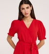 A woman with long dark hair wears the Everleigh Red Crepe Midi Dress, a red short-sleeve midi dress with a faux wrap neckline and waist tie, standing against a plain light background.