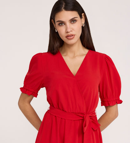 A woman with long dark hair wears the Everleigh Red Crepe Midi Dress, a red short-sleeve midi dress with a faux wrap neckline and waist tie, standing against a plain light background.