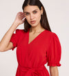 A woman with long brown hair wears the Everleigh Red Crepe Midi Dress, featuring a faux wrap neckline, puff sleeves, and waist tie, posing with one hand near her ear and looking at the camera against a light background.