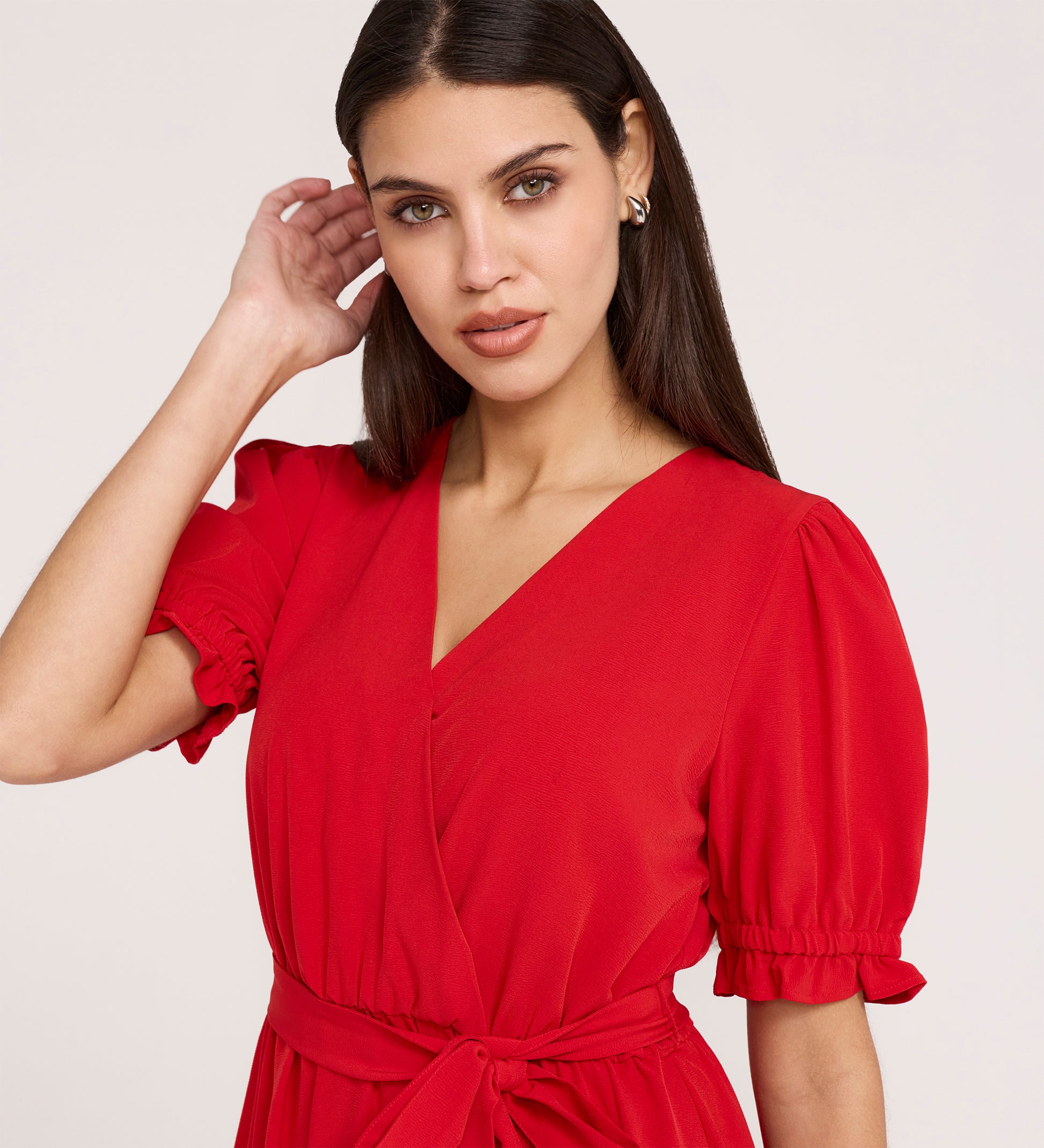 A woman with long brown hair wears the Everleigh Red Crepe Midi Dress, featuring a faux wrap neckline, puff sleeves, and waist tie, posing with one hand near her ear and looking at the camera against a light background.