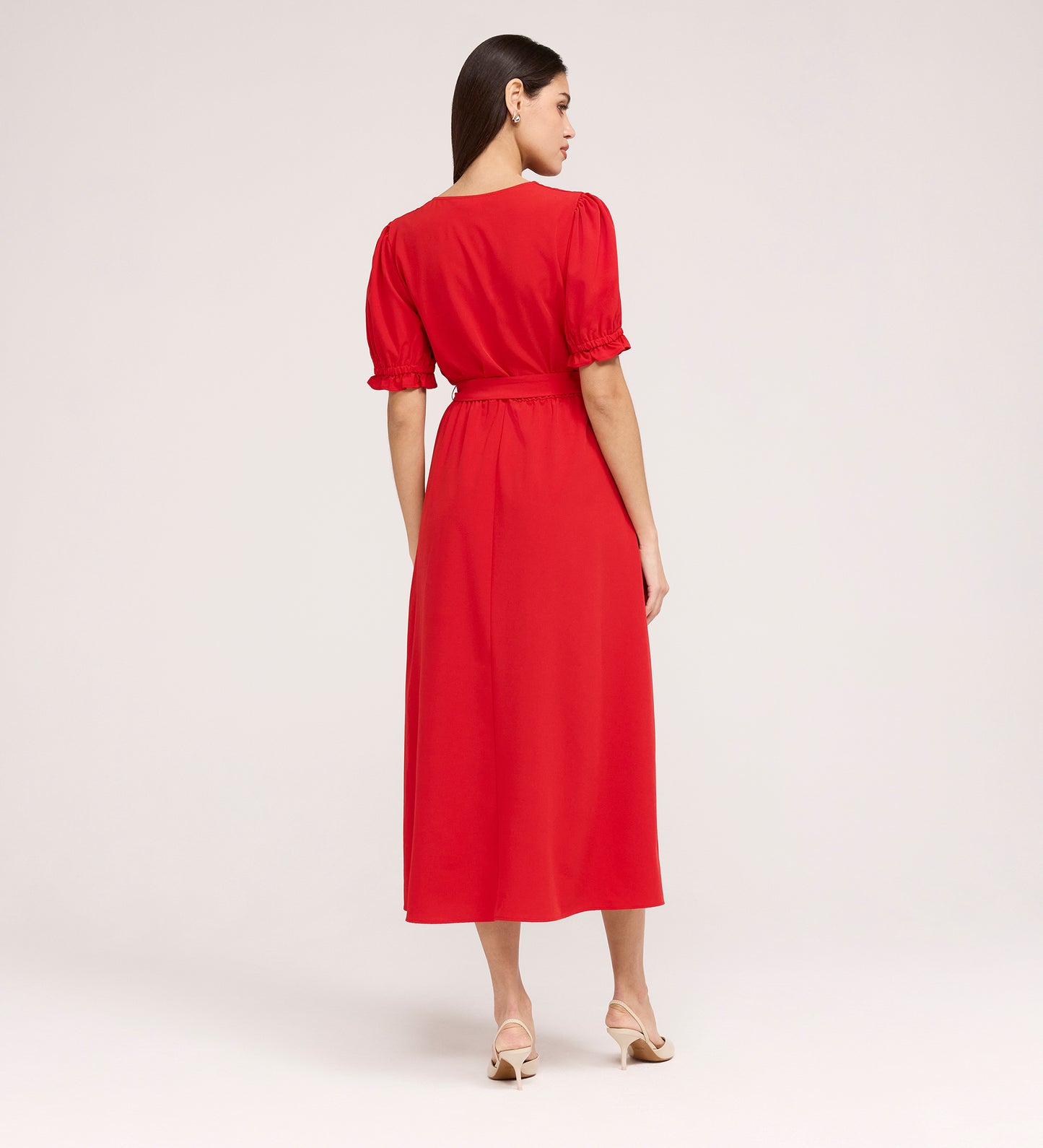 A woman with straight dark hair, in beige heeled sandals, faces away wearing the Everleigh Red Crepe Midi Dress—a vibrant red midi with puffed short sleeves and a gathered waist—set against a plain light background.