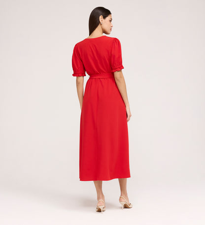 A woman with straight dark hair, in beige heeled sandals, faces away wearing the Everleigh Red Crepe Midi Dress—a vibrant red midi with puffed short sleeves and a gathered waist—set against a plain light background.