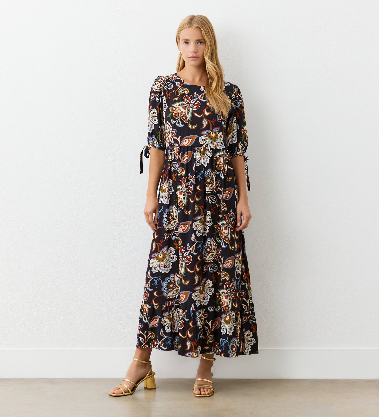 A woman with long blonde hair stands against a white wall, wearing the Gwen Black Floral Midi Smock Dress featuring a relaxed fit, puff sleeves, and a tiered hem, paired with gold strappy sandals.