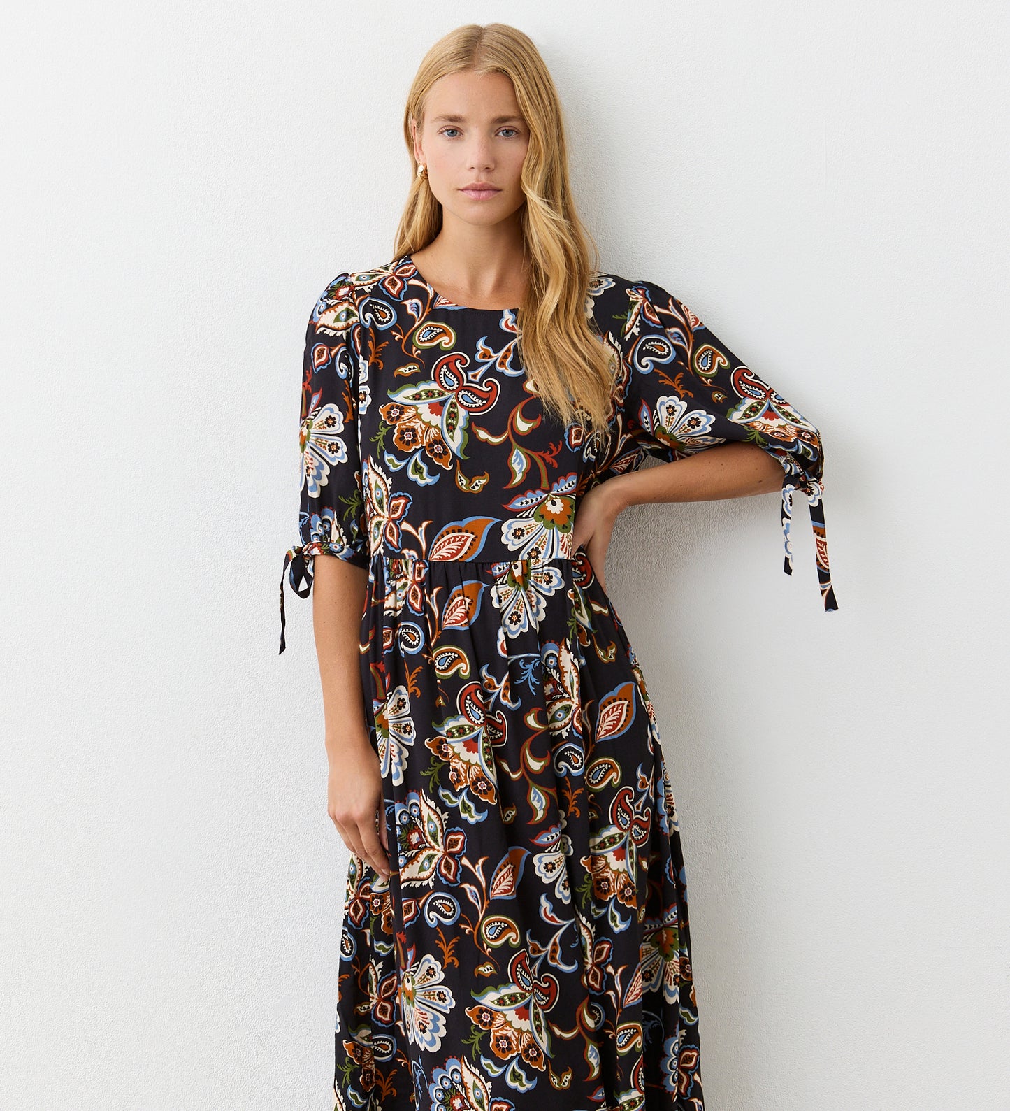 A woman with long blonde hair wears the Gwen Black Floral Midi Smock Dress, a relaxed fit black midi dress with a vibrant floral pattern and short tie sleeves, standing against a plain white wall.