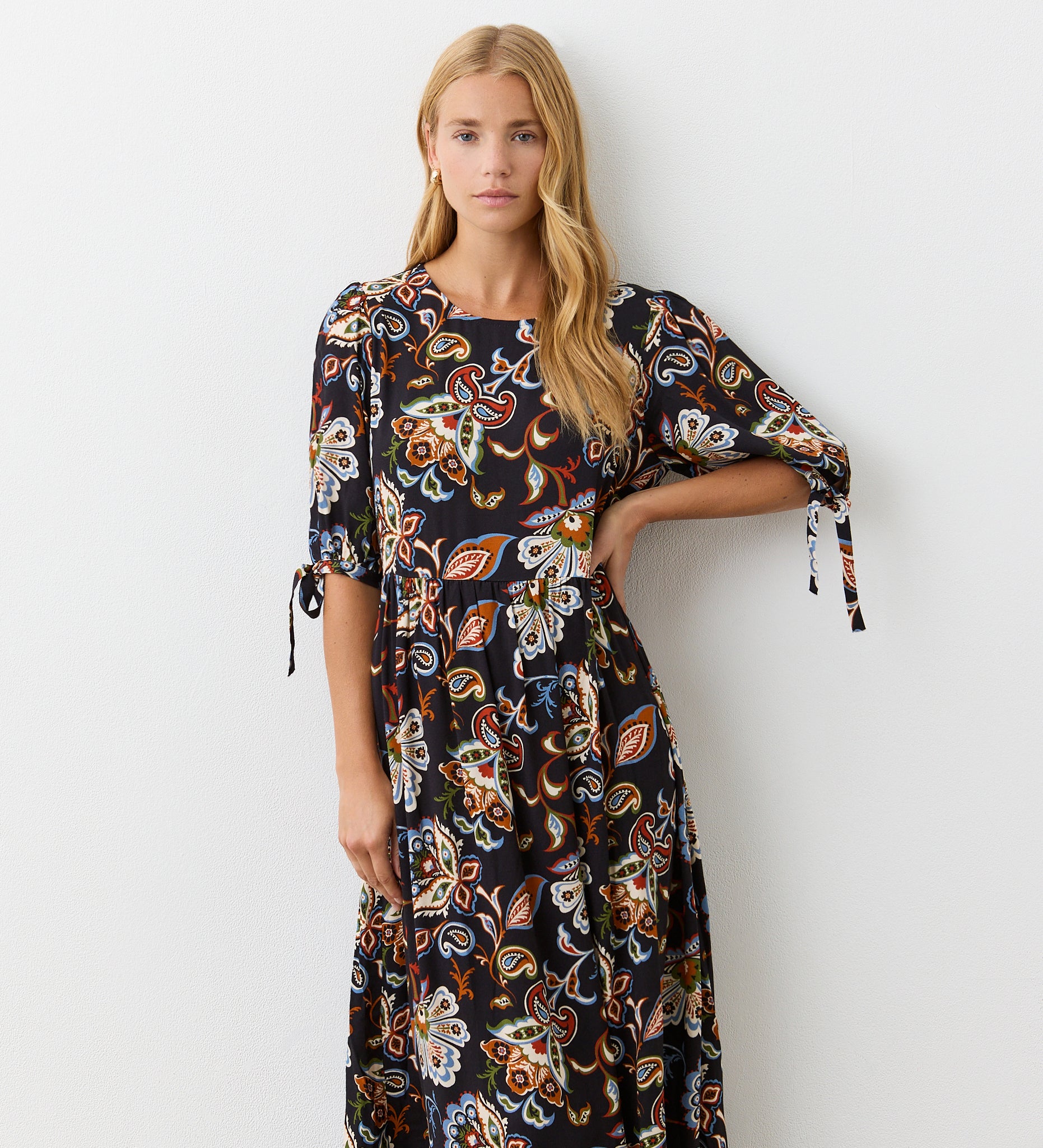A woman with long blonde hair wears the Gwen Black Floral Midi Smock Dress, a relaxed fit black midi dress with a vibrant floral pattern and short tie sleeves, standing against a plain white wall.