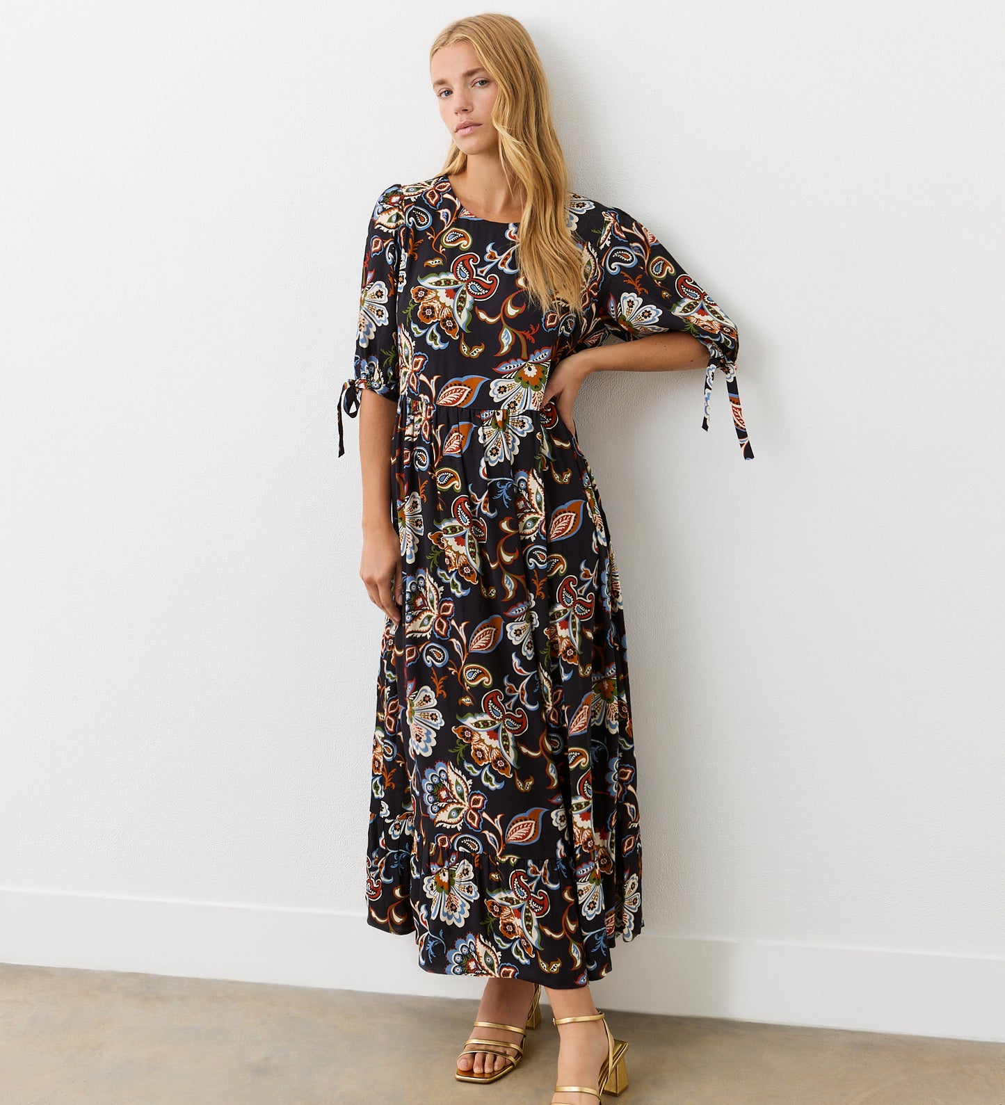 A woman stands against a white wall wearing the Gwen Black Floral Midi Smock Dress, featuring a vibrant floral and paisley print, relaxed fit, tiered hem, and styled with gold sandals. She has long blonde hair and one hand on her hip.