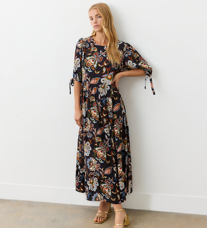 A woman stands against a white wall wearing the Gwen Black Floral Midi Smock Dress, featuring a vibrant floral and paisley print, relaxed fit, tiered hem, and styled with gold sandals. She has long blonde hair and one hand on her hip.