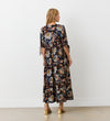 A woman with blonde hair faces a white wall, wearing the Gwen Black Floral Midi Smock Dress featuring a relaxed fit, colorful floral and paisley pattern, tiered hem, short tied sleeves, and gold sandals.
