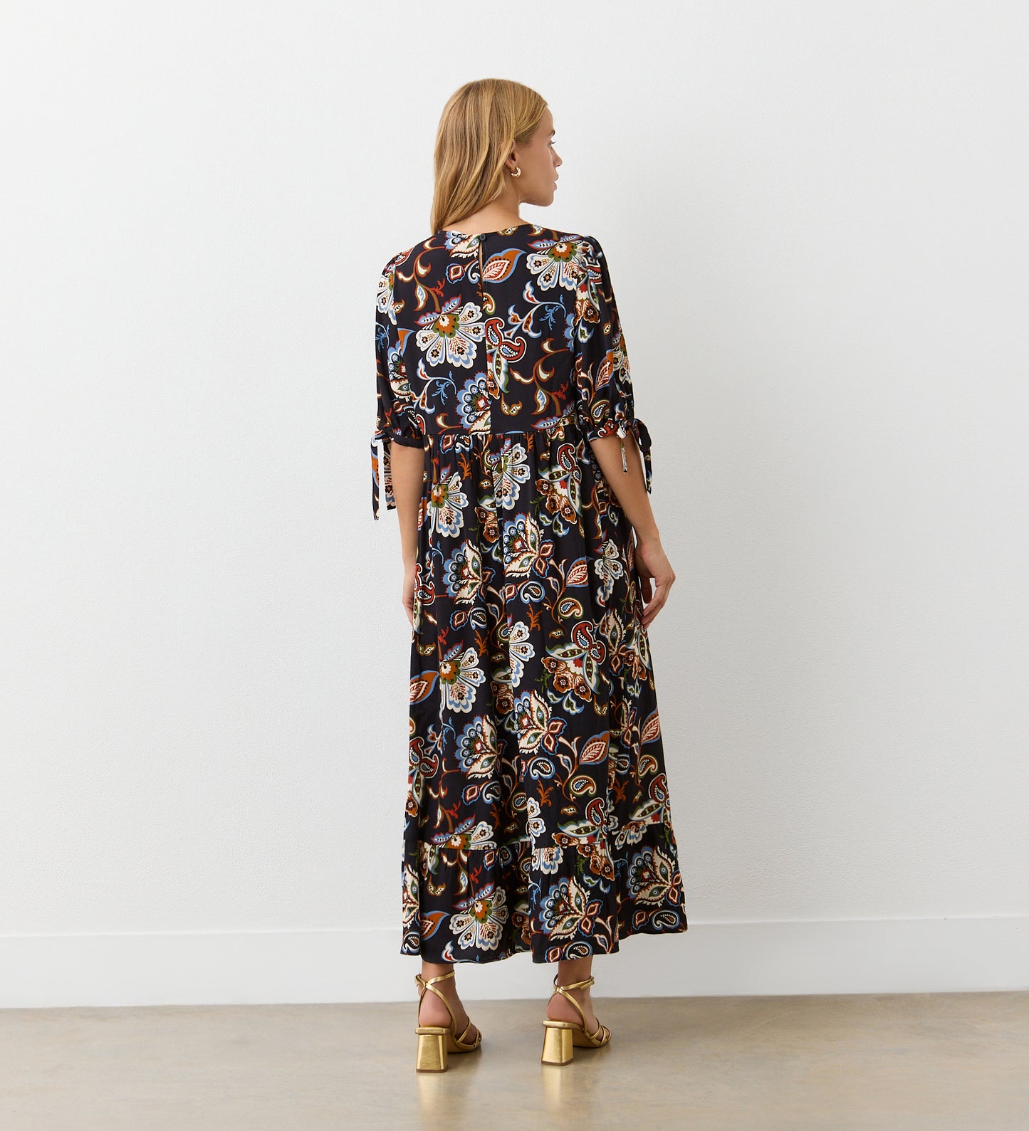 A woman with blonde hair faces a white wall, wearing the Gwen Black Floral Midi Smock Dress featuring a relaxed fit, colorful floral and paisley pattern, tiered hem, short tied sleeves, and gold sandals.