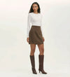 A woman with long curly hair wears a fitted white long-sleeve top, the Helia Mole Ponte Jersey Mini Skirt with an elasticated waistband, knee-high brown boots, and a simple necklace against a plain background.