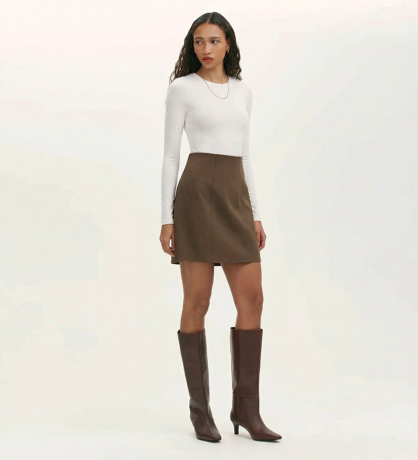 A woman with long curly hair wears a fitted white long-sleeve top, the Helia Mole Ponte Jersey Mini Skirt with an elasticated waistband, knee-high brown boots, and a simple necklace against a plain background.