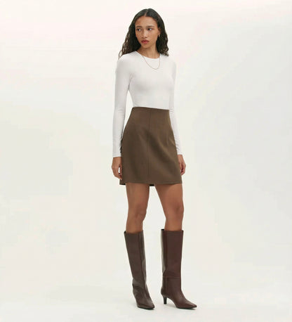 A woman with long curly hair wears a fitted white long-sleeve top, the Helia Mole Ponte Jersey Mini Skirt with an elasticated waistband, knee-high brown boots, and a simple necklace against a plain background.