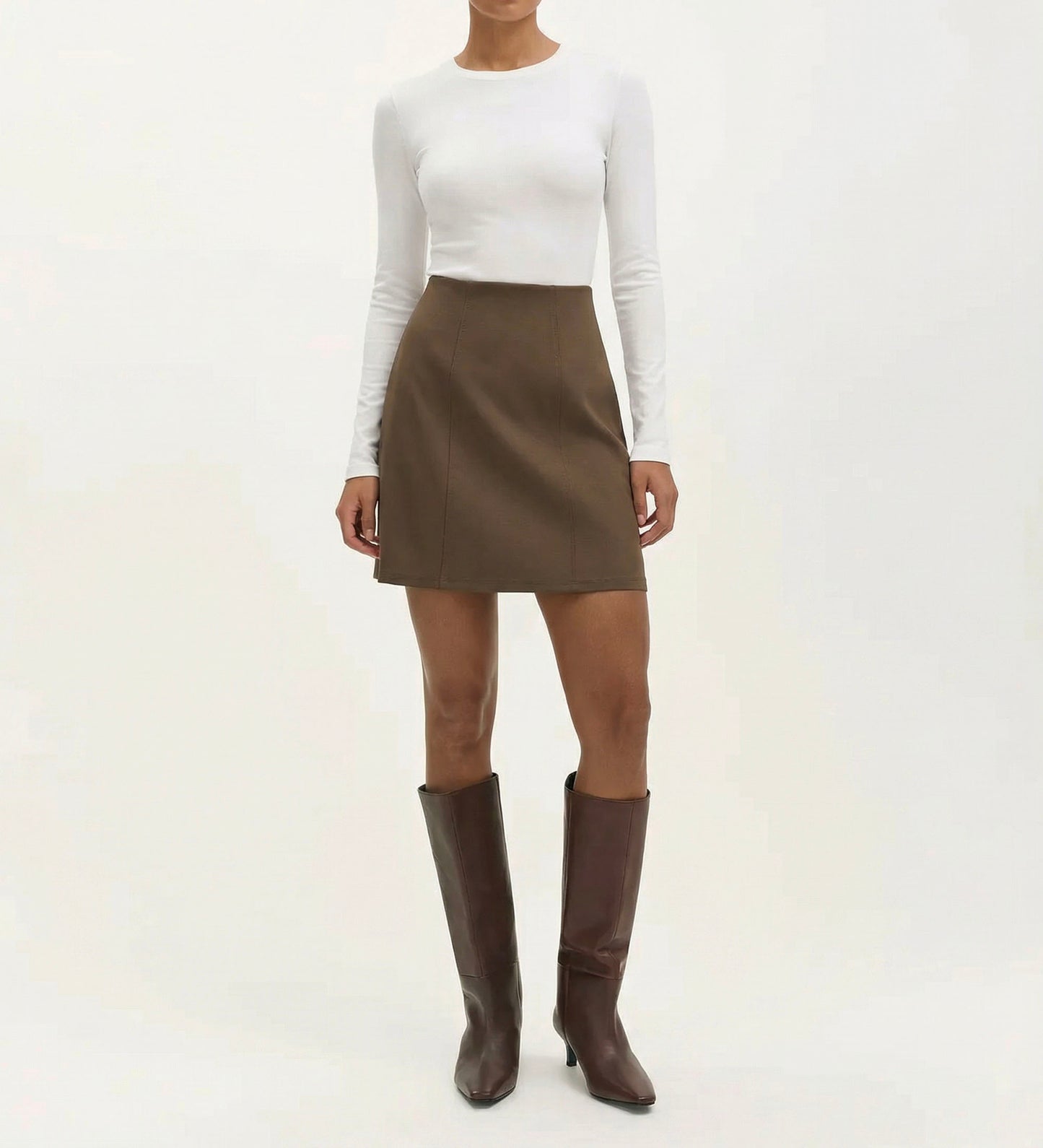 Someone stands against a plain light background wearing a fitted white long-sleeve top, the Helia Mole Ponte Jersey Mini Skirt featuring an elasticated waistband, and tall brown heeled boots.