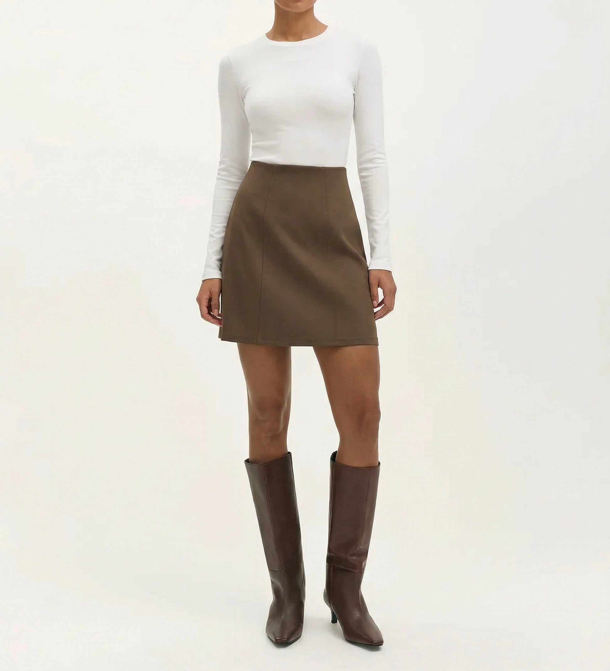 Someone stands against a plain light background wearing a fitted white long-sleeve top, the Helia Mole Ponte Jersey Mini Skirt featuring an elasticated waistband, and tall brown heeled boots.