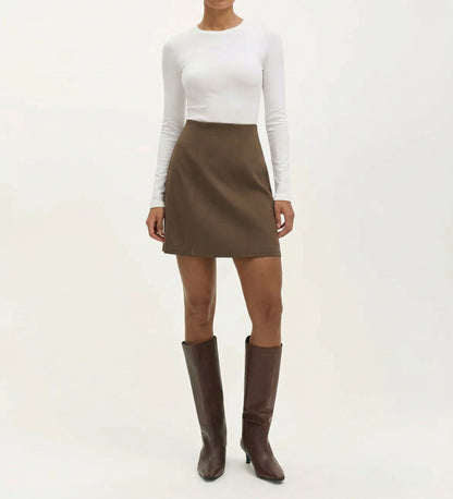 Someone stands against a plain light background wearing a fitted white long-sleeve top, the Helia Mole Ponte Jersey Mini Skirt featuring an elasticated waistband, and tall brown heeled boots.