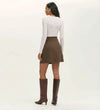 A woman faces away, wearing a fitted long-sleeve white top, knee-high brown boots, and the Helia Mole Ponte Jersey Mini Skirt—high-waisted with an elasticated waistband—set against a plain light background.