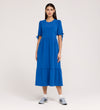 A woman stands against a plain background, wearing the Jessa Cobalt Crepe Midi Tiered Dress with puff sleeves, sleeve bows, and a tiered skirt, paired with gray athletic sneakers.