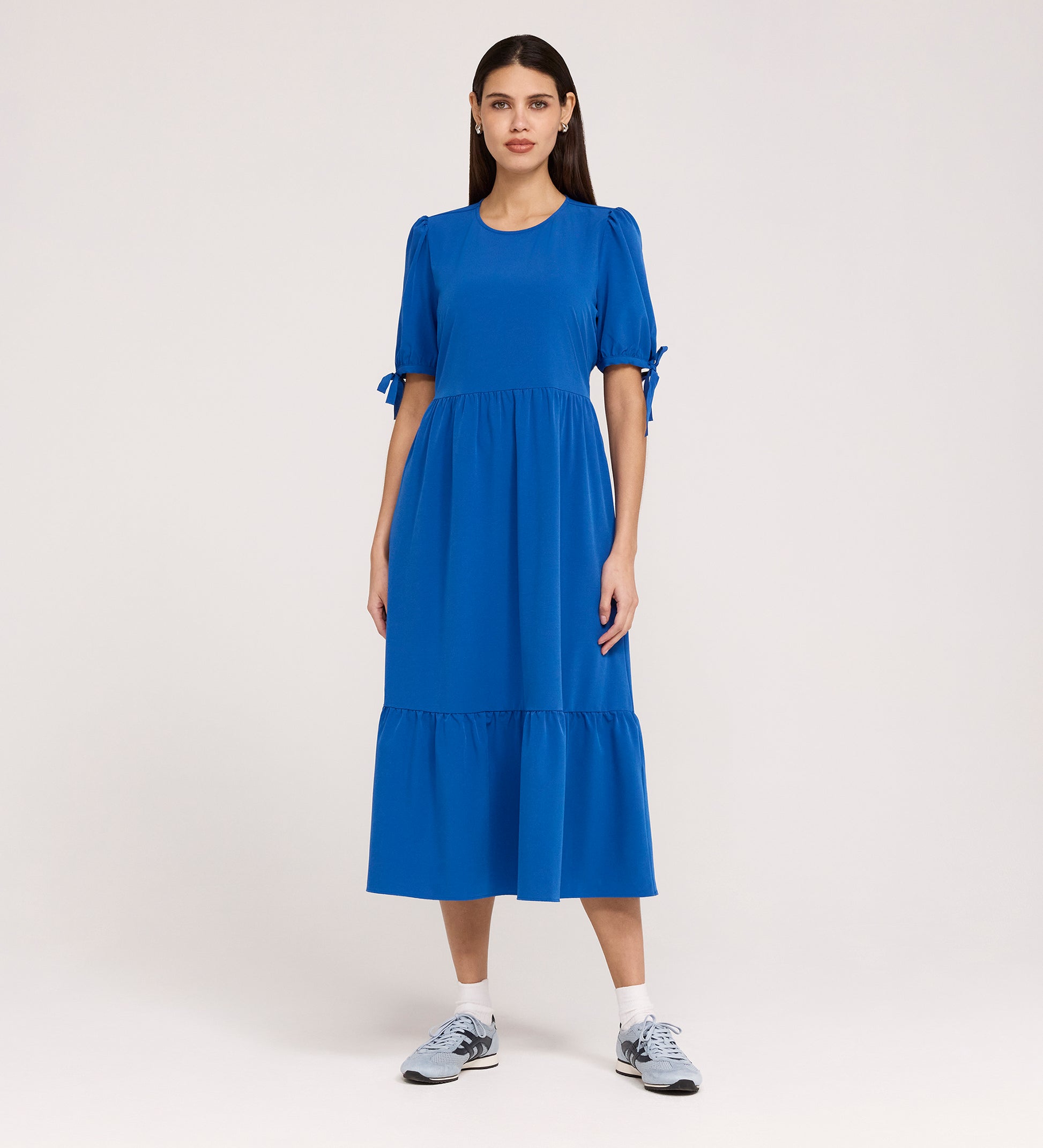 A woman stands against a plain background, wearing the Jessa Cobalt Crepe Midi Tiered Dress with puff sleeves, sleeve bows, and a tiered skirt, paired with gray athletic sneakers.