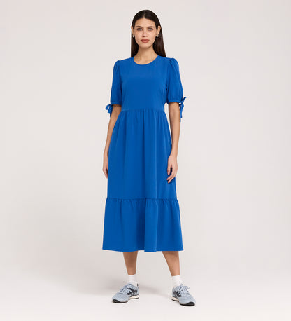 A woman stands against a plain background, wearing the Jessa Cobalt Crepe Midi Tiered Dress with puff sleeves, sleeve bows, and a tiered skirt, paired with gray athletic sneakers.
