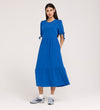 A woman poses against a plain light background in the Jessa Cobalt Crepe Midi Tiered Dress, featuring tie details on puff sleeves and a ruffled, tiered skirt, styled with gray sneakers.