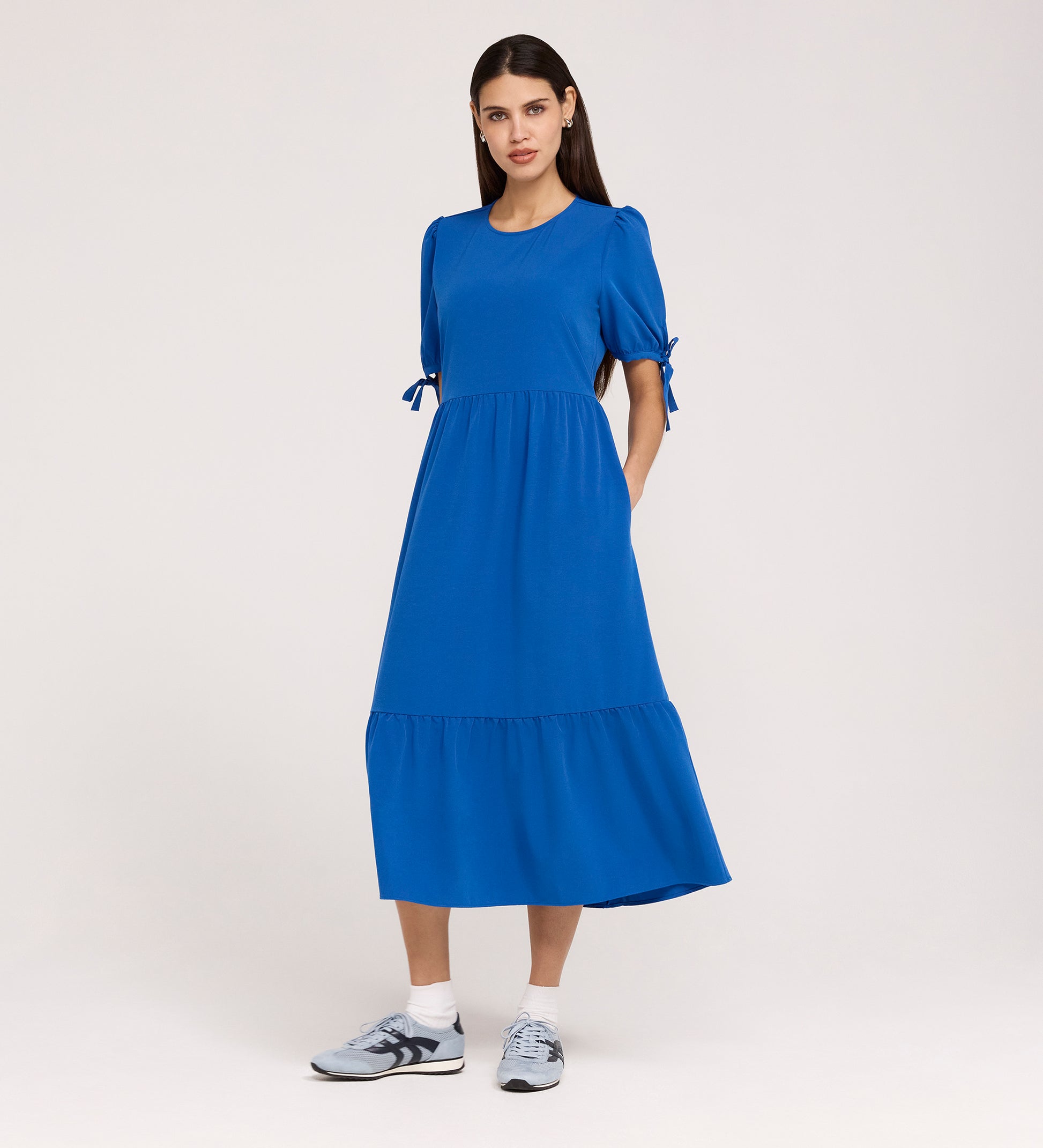 A woman poses against a plain light background in the Jessa Cobalt Crepe Midi Tiered Dress, featuring tie details on puff sleeves and a ruffled, tiered skirt, styled with gray sneakers.