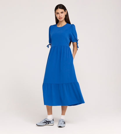 A woman poses against a plain light background in the Jessa Cobalt Crepe Midi Tiered Dress, featuring tie details on puff sleeves and a ruffled, tiered skirt, styled with gray sneakers.