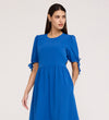 A woman with long dark hair wears the Jessa Cobalt Crepe Midi Tiered Dress, which has short puff sleeves with bows and a tiered skirt, standing against a plain light background and looking to the side.