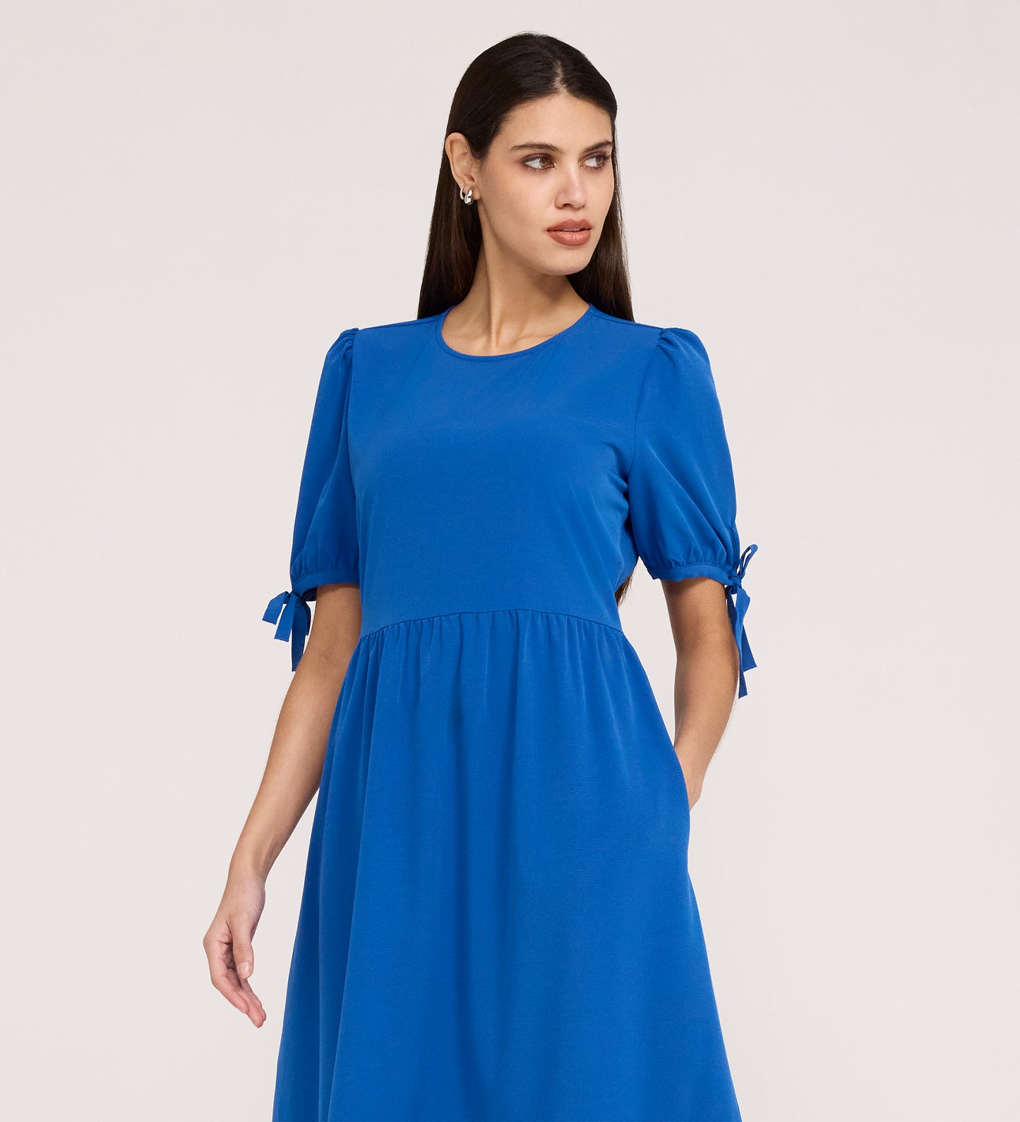 A woman with long dark hair wears the Jessa Cobalt Crepe Midi Tiered Dress, which has short puff sleeves with bows and a tiered skirt, standing against a plain light background and looking to the side.