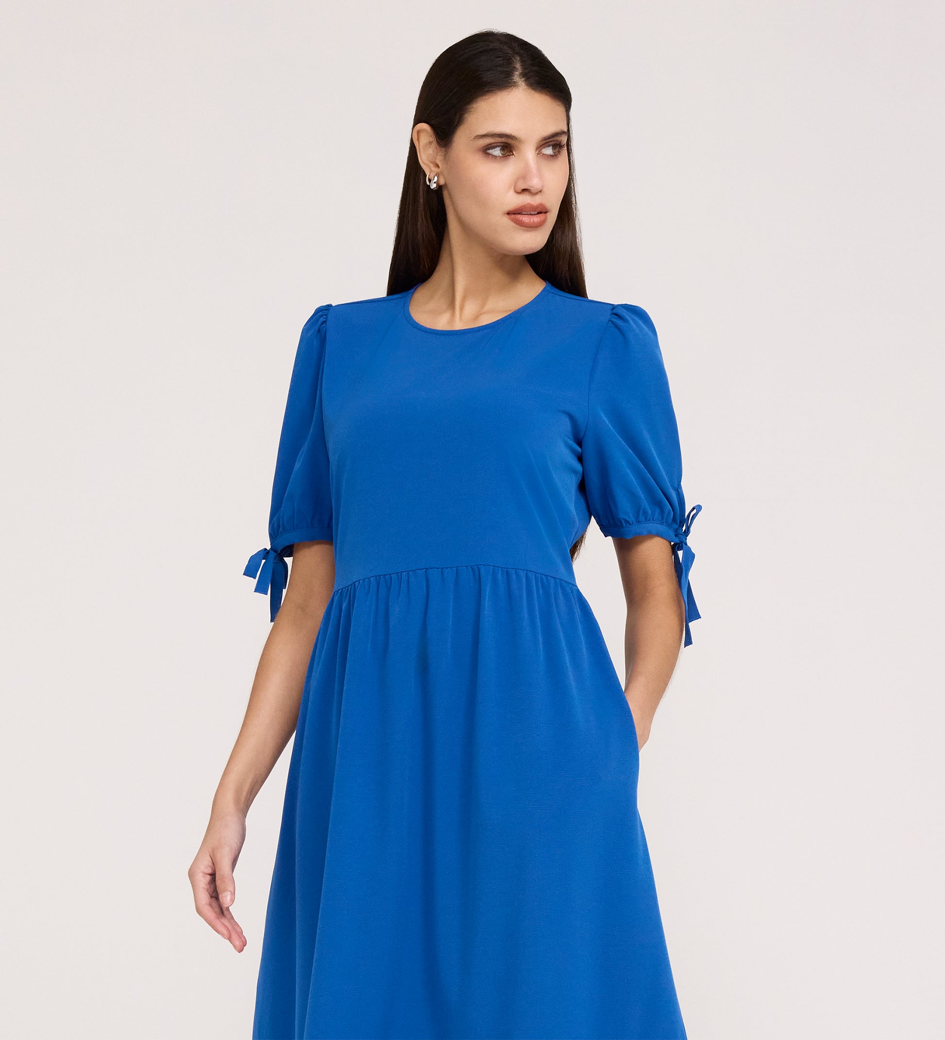 A woman with long dark hair wears the Jessa Cobalt Crepe Midi Tiered Dress, which has short puff sleeves with bows and a tiered skirt, standing against a plain light background and looking to the side.