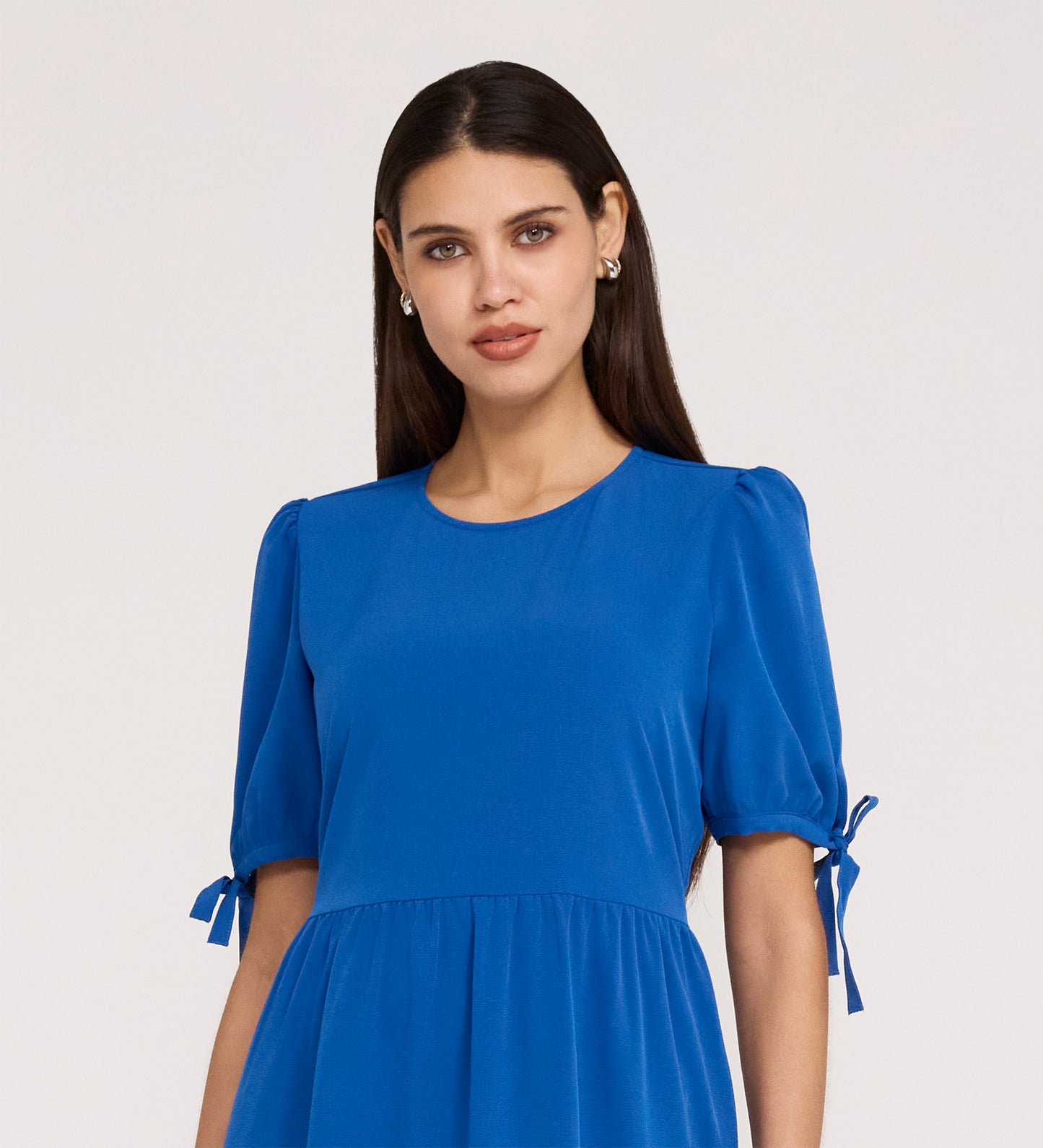 A woman with long dark hair models the Jessa Cobalt Crepe Midi Tiered Dress, featuring short puff sleeves and bow details. She stands against a plain light background, looking at the camera with a neutral expression.
