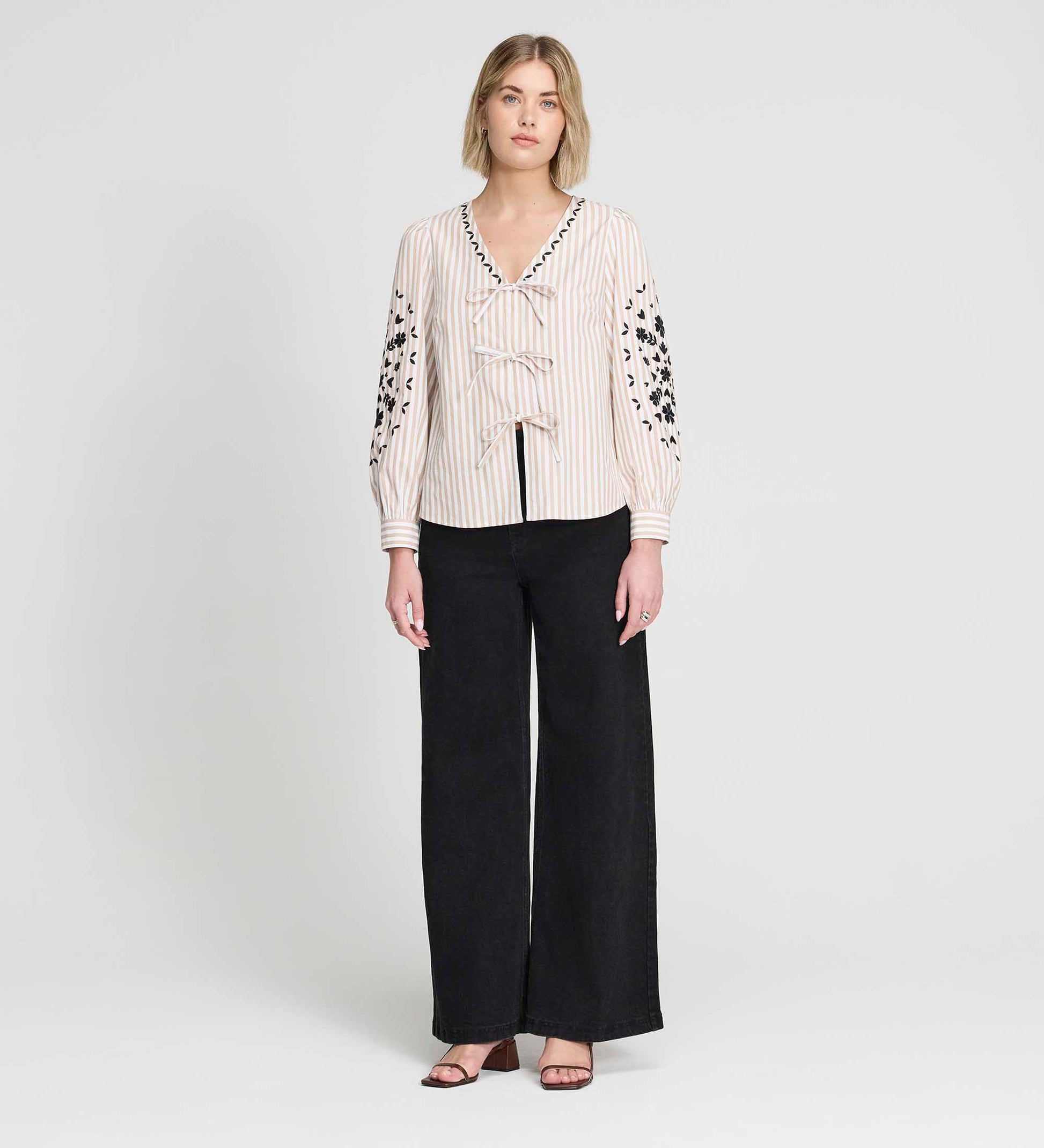 Woman wearing Karlee Neutral Stripe Embroidered Cotton Tie Front Blouse and black pants on a white background