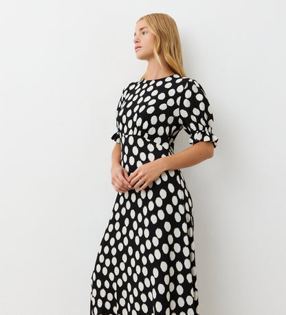 A woman with long, straight blonde hair stands against a white wall, wearing the Mela Black Polka Dot Crepe Midi Dress with short sleeves and looking calmly to the side.