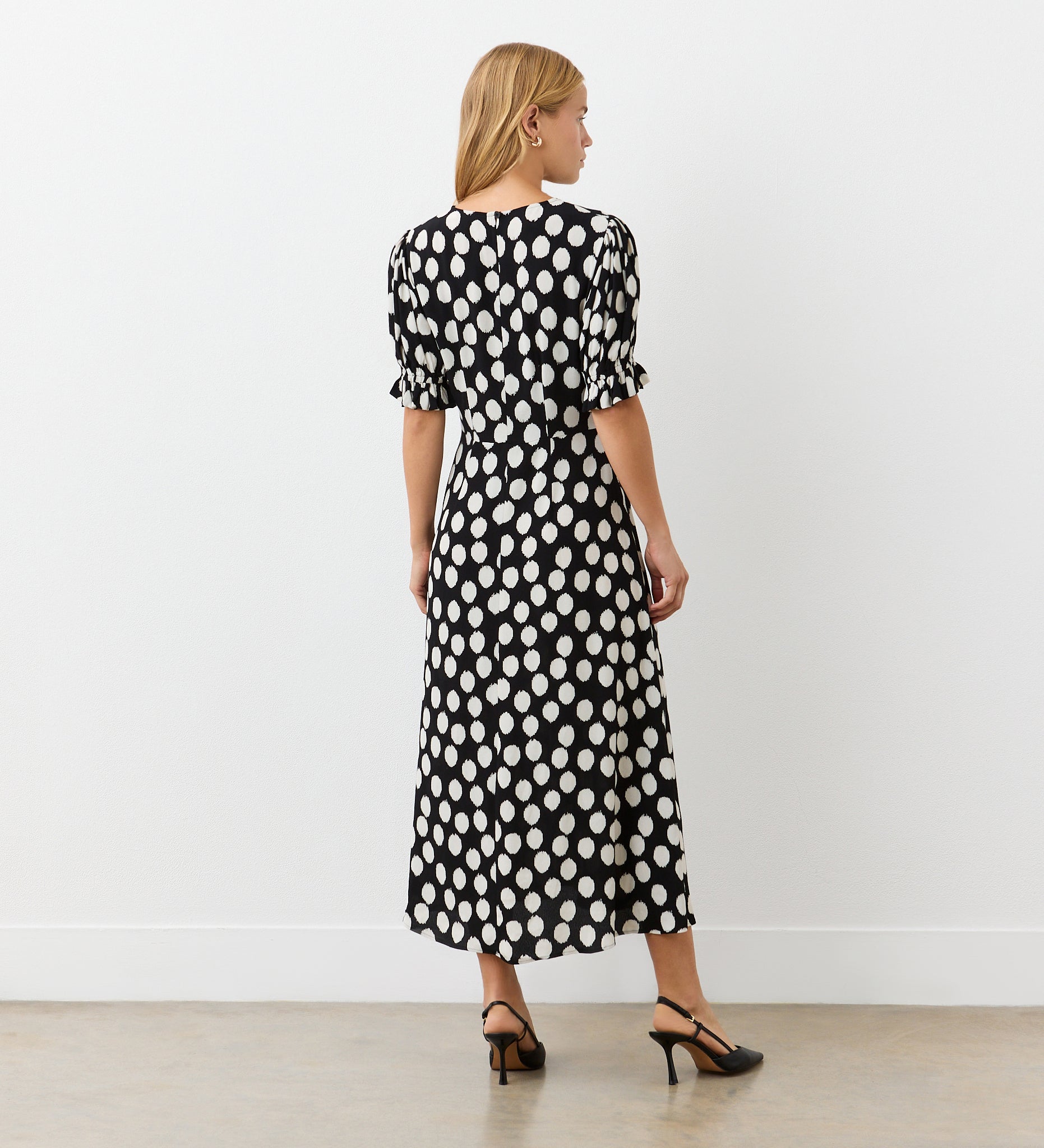 A woman with blonde hair faces away, wearing the Mela Black Polka Dot Crepe Midi Dress with large white dots, short puff sleeves, and black heels, set against a plain white wall and light-colored floor.