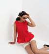 Woman wearing a Malani Red Cotton Poplin Shirred Peplum Shell Top and white shorts on a white background